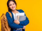 Happy asian female college student smiling at camera on yellow background and copy space, holding laptop and books, hanging bagpack. Youth girl student is exchange student. education concept